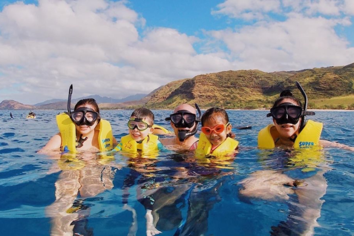 Five snorkelers with life vests in clear blue water near mountains.