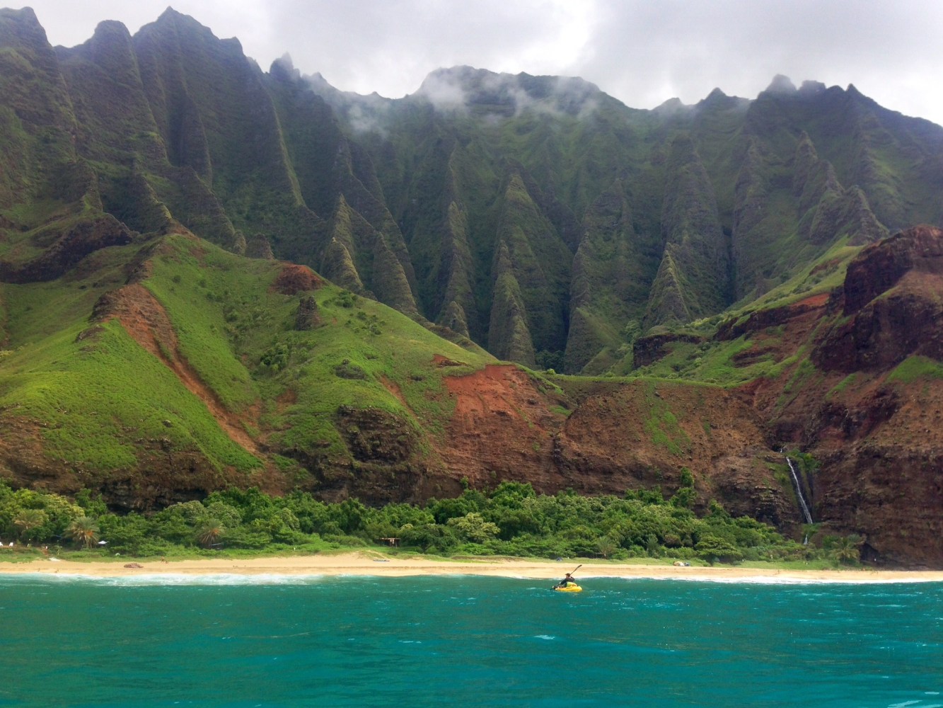 Lush green cliffs with clouds above, beach, and blue ocean with a kayaker.