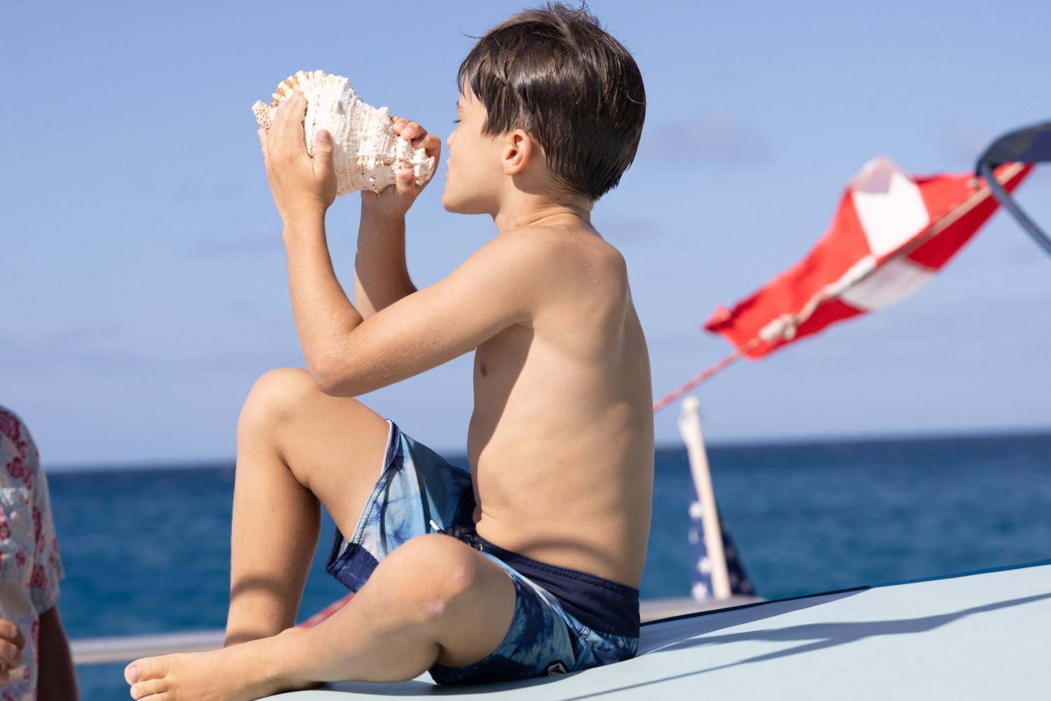 Boy on a boat holding a large seashell to his mouth, ocean in background.
