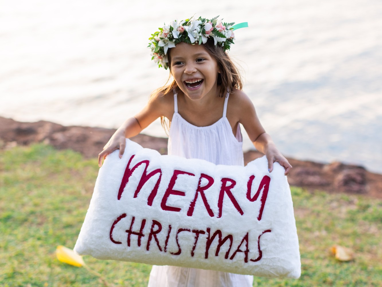 Girl in white dress and flower crown holds 'Merry Christmas' pillow outside.