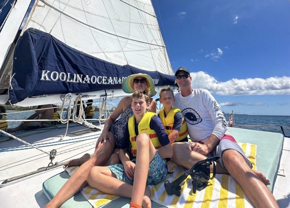 Family of four on a sailboat, under a sunlit sky, wearing life vests and hats.