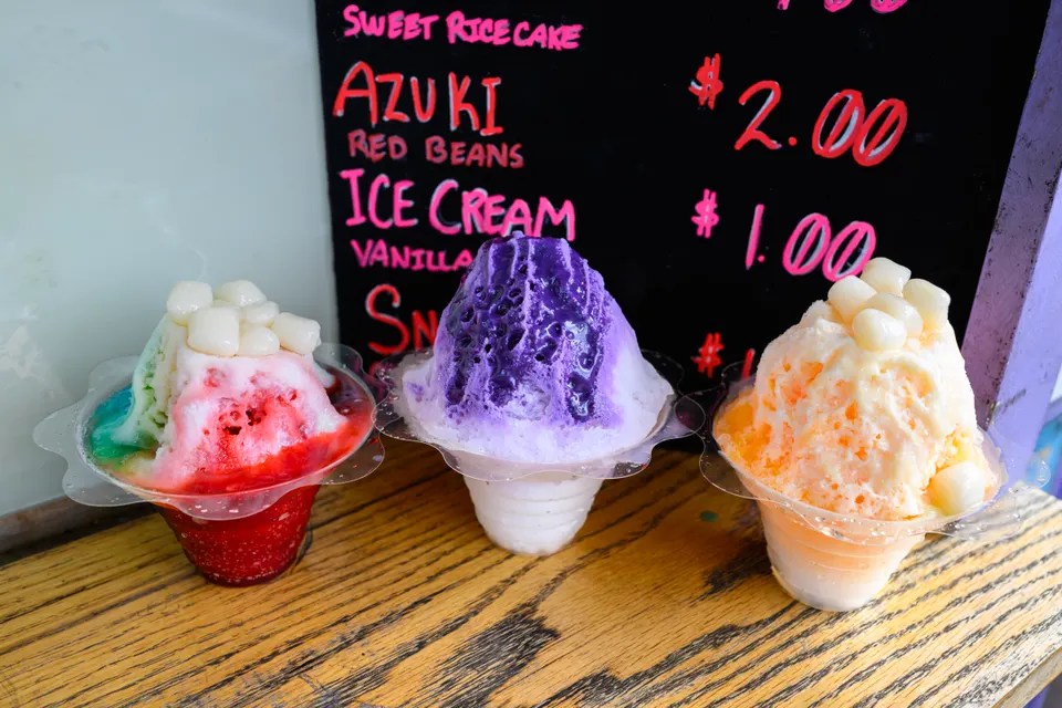 Three colorful shaved ice desserts in cups on a wood table next to a menu.