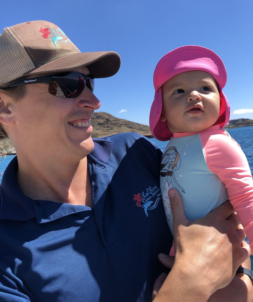 Adult holding a baby wearing a pink hat and swimsuit, with a sunny outdoor backdrop.