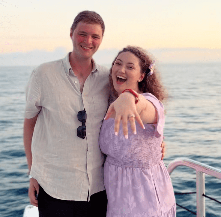 Happy couple on a boat; woman shows ring, ocean at sunset.