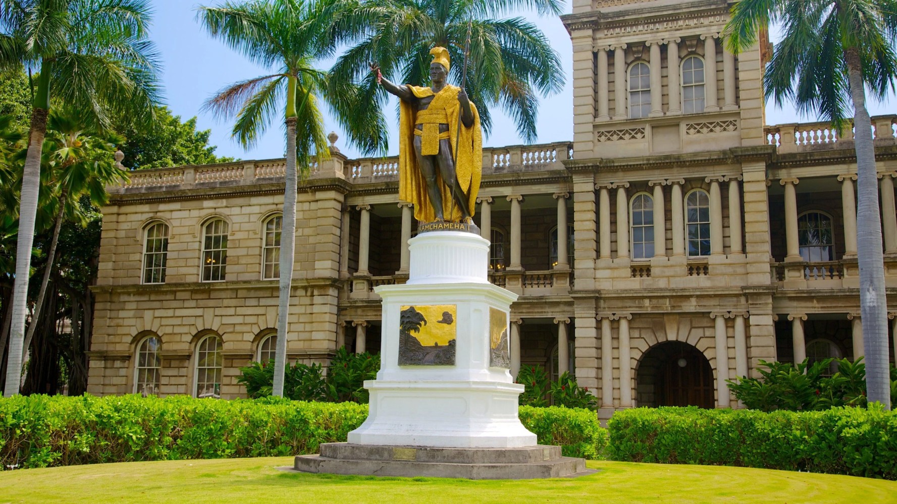 Statue of a figure in gold cloak in front of a historic building with palm trees.